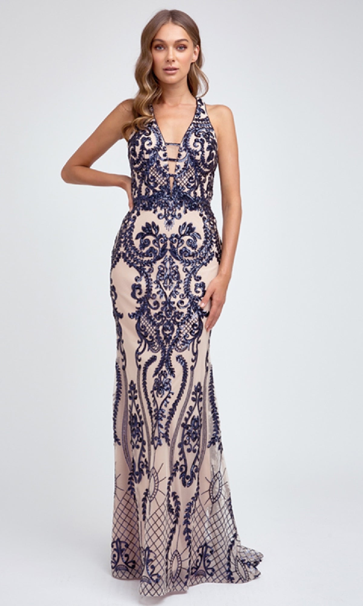 A woman models the Juliet 243 Long Prom Dress, a sleeveless, floor-length gown with intricate navy blue embroidery over nude fabric, set against a plain light background.